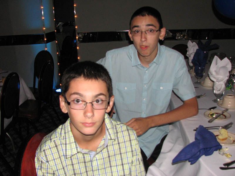 Two young people at a table, one seated wearing glasses and a plaid shirt, the other standing, wearing a light blue shirt.