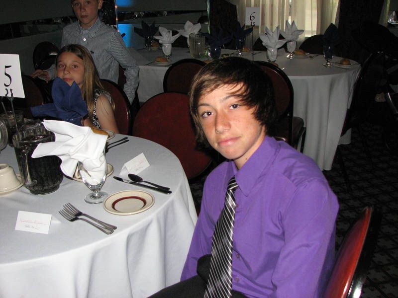 Teen in purple shirt and striped tie seated at table set for a formal event.