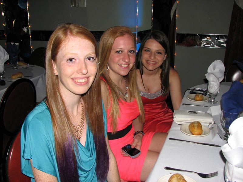 Three young women smiling at a table, likely at a formal event. One in teal dress, one in coral, one in red.