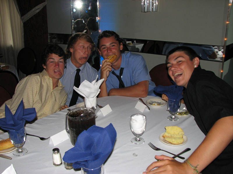 Four young people at a table; one eating, others smiling. Blue napkins, white tablecloth, formal setting.