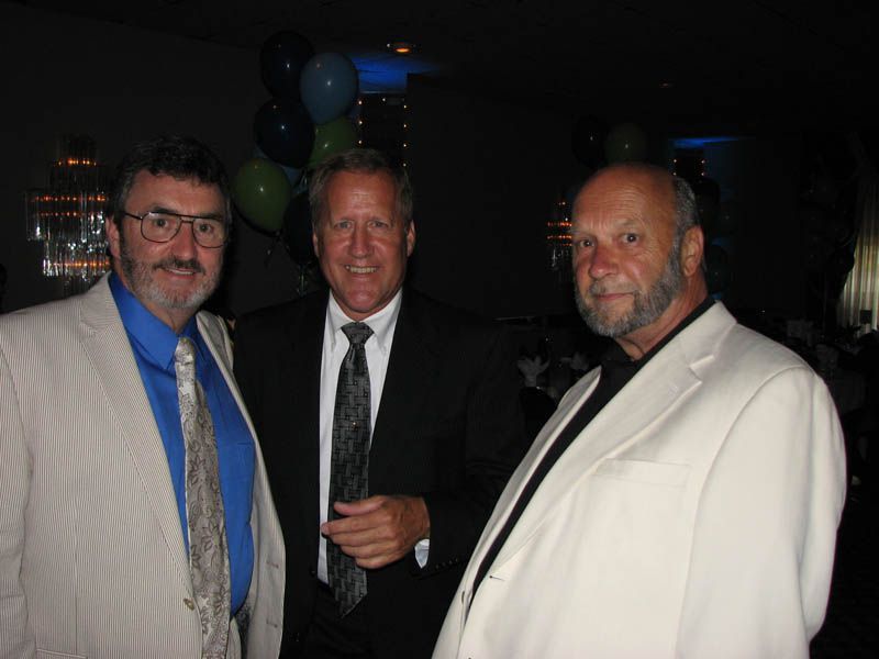 Three men in suits pose indoors. Background has balloons.