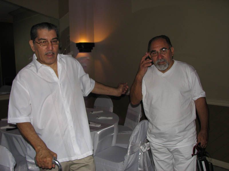Two men in white shirts, one with a cane and arm raised, the other on the phone, inside a room with tables.
