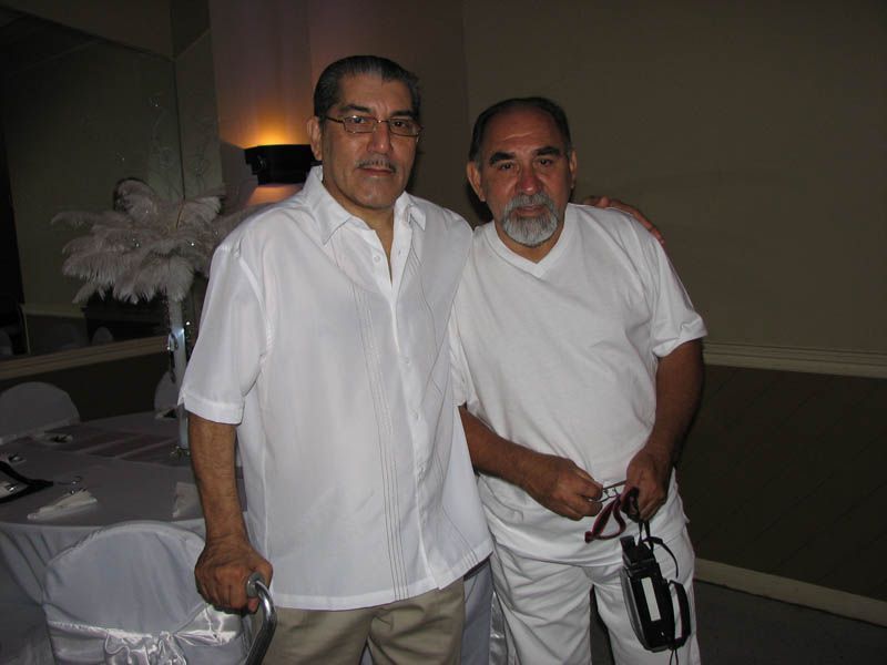 Two men pose together indoors. Both are wearing white. One man has a beard and holds a video camera.