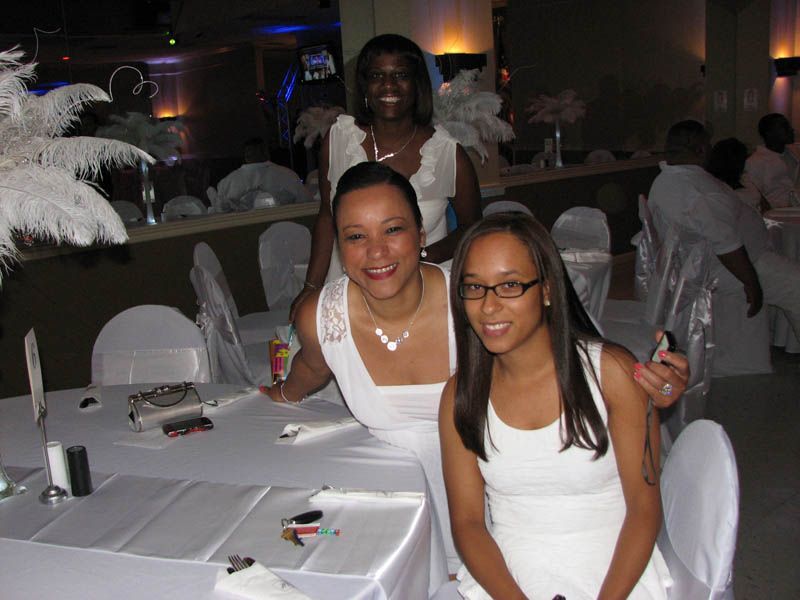 Three people in white outfits pose at a table. Decorative feathers and other guests are in the background.