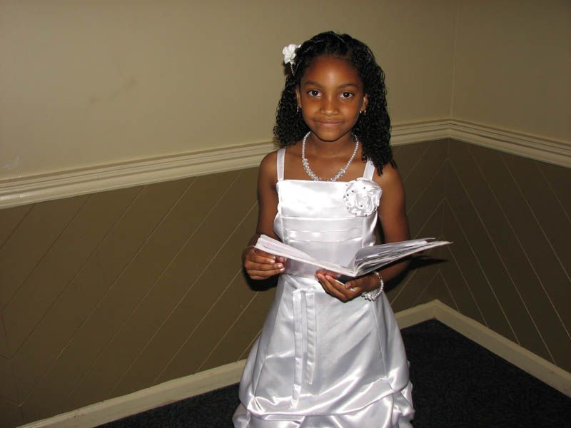 Girl in white dress holding papers, standing against a tan wall.