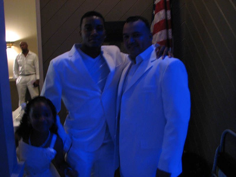 Three people in white suits pose for a photo with a young girl in a white dress, indoors with blue lighting.