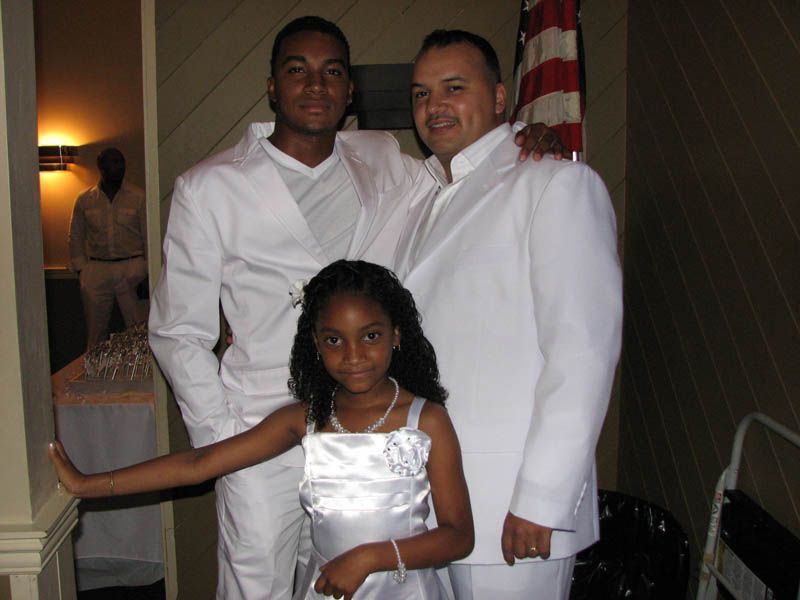 Three people in white formal wear pose indoors: a young girl, a man, and a young adult.