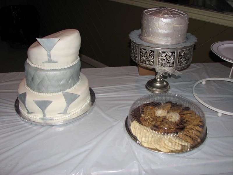 Three-tiered cake with martini glasses, a small silver cake, and a cookie platter on a white table.