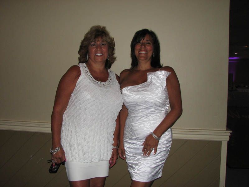 Two women in white dresses smiling in front of a neutral wall.