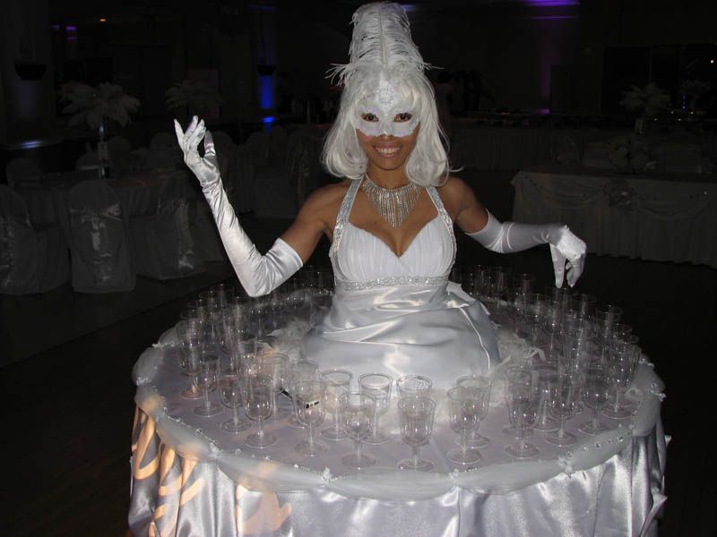 Woman in white dress with mask, serving champagne glasses on a moving table.