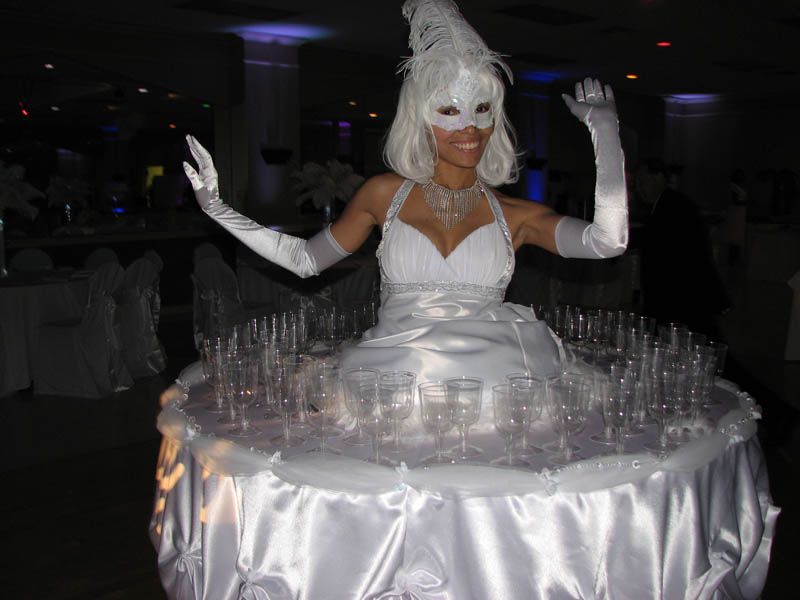 Woman in white dress with champagne glasses on a table skirt, wearing a mask and gloves, at an event.