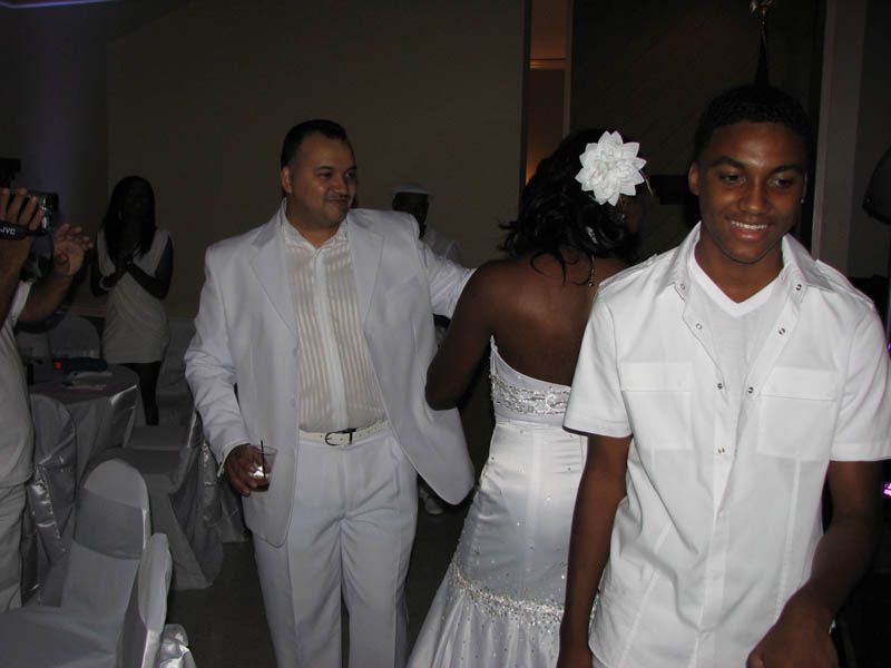 People in white attire at an event, including a bride in a white dress, a man in a suit, and a man in a button-down.