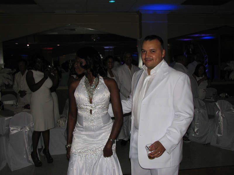 Couple in white formal wear at an event; woman with beaded dress, man with suit, others in background.
