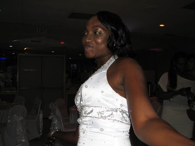Woman in white halter dress smiles, arms extended. Inside venue with lights and tables.