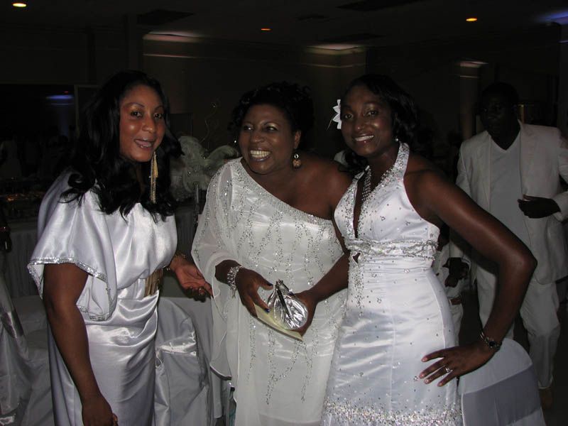 Three women smile in white formal wear at an indoor event, with a man blurred in the background.