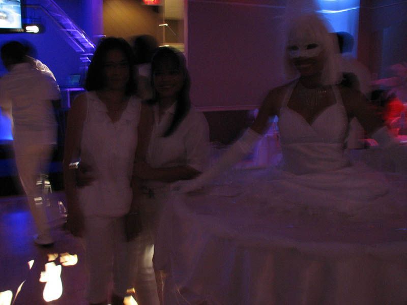 Two women in white outfits pose near a masked performer in white. Party setting with blue and pink lighting.