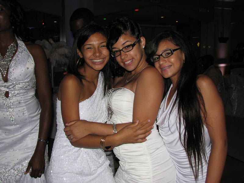 Three smiling people in white dresses hug at a party.