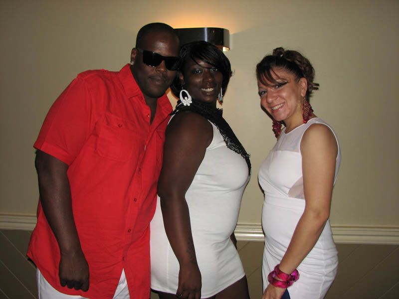 Three people pose indoors: man in red shirt, woman in white dress, woman in pink and white dress.