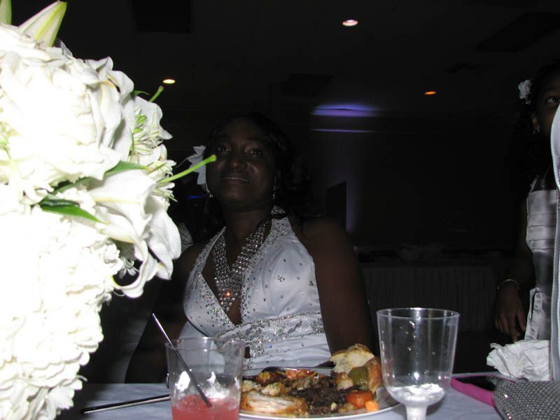 Woman in a white dress smiles at a table with flowers, food, and drinks in a dimly lit room.
