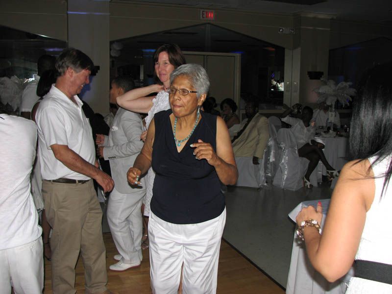 Elderly person dancing at a party, wearing white pants and a navy top. Other people are in the background.