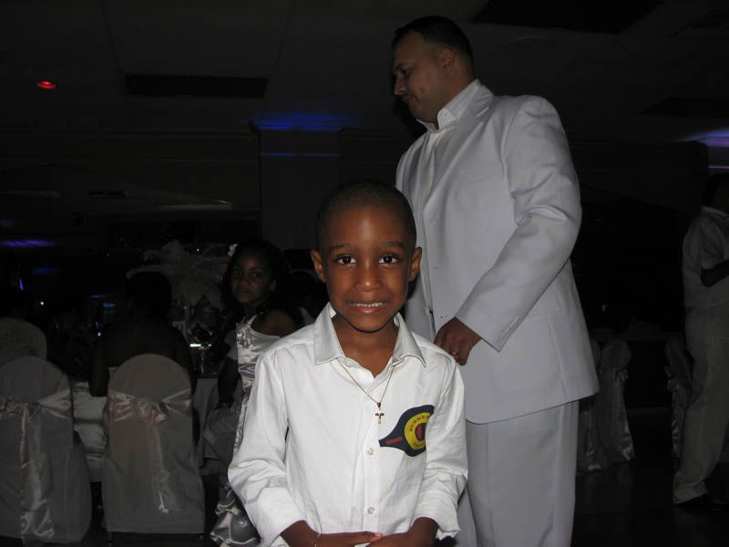 Boy in white shirt smiles, standing in a dim room. Man in white suit stands behind him.