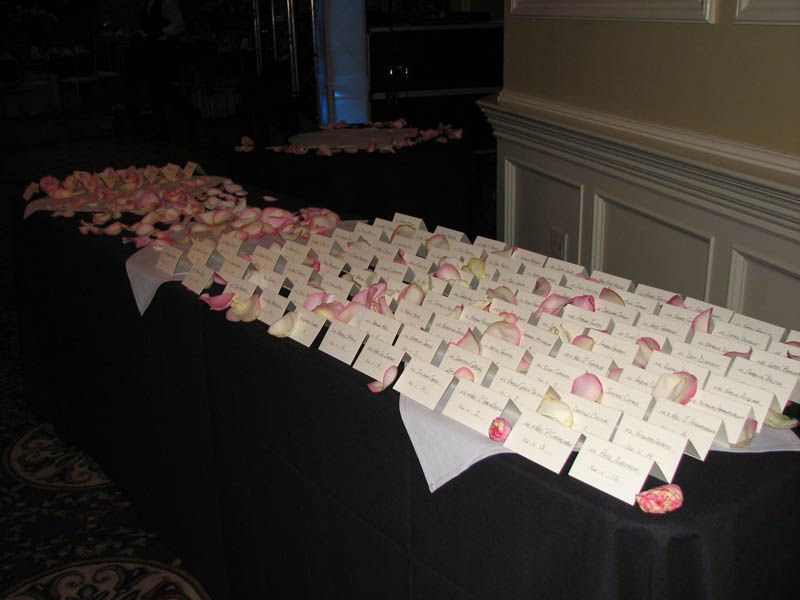 Table with place cards and scattered pink rose petals. Black tablecloth, neutral wall in background.