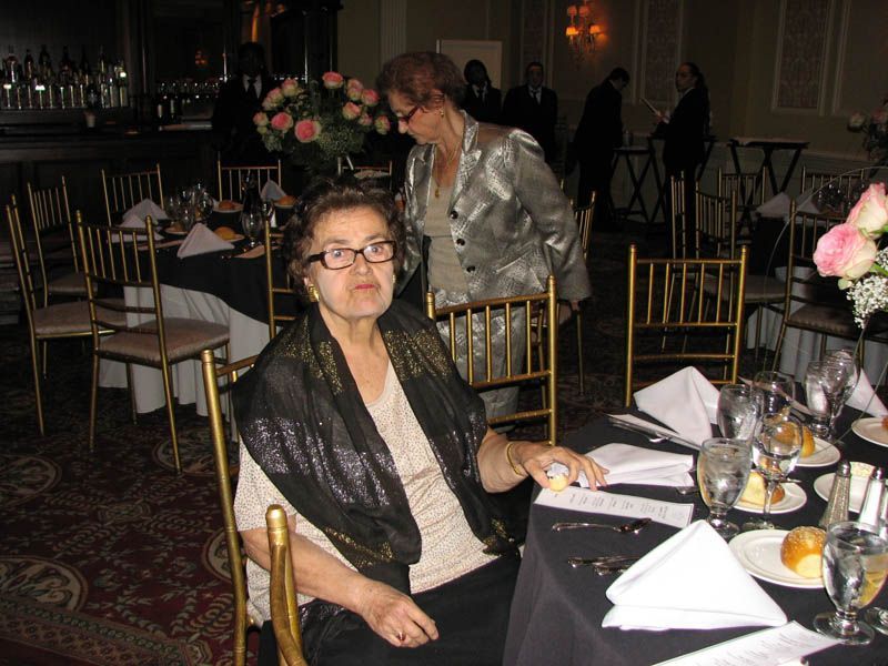 Woman at a table in a formal setting, wearing glasses and shawl, another woman standing behind her.