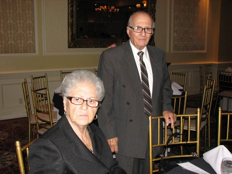 Elderly couple at a formal event; woman seated, man standing, both wearing glasses and suits.