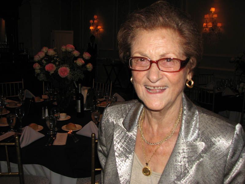 Woman in silver jacket, glasses, and gold jewelry smiling at a formal event with flower centerpieces.