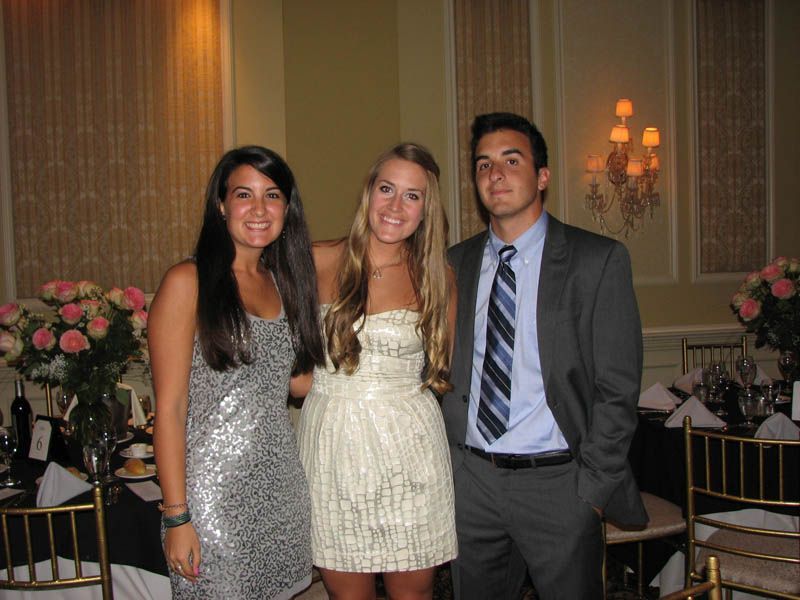 Three people at an event pose together. Woman in silver dress, woman in white dress, man in gray suit. Floral backdrop.