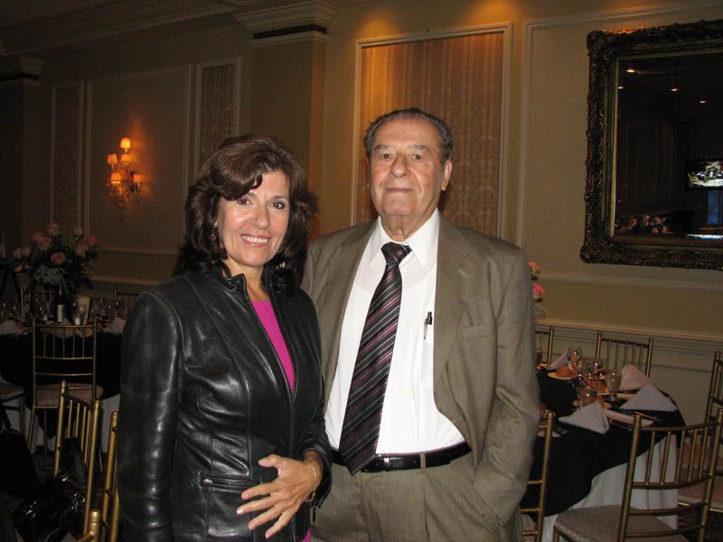 Woman in black jacket and man in suit, smiling, standing indoors near a dining table.