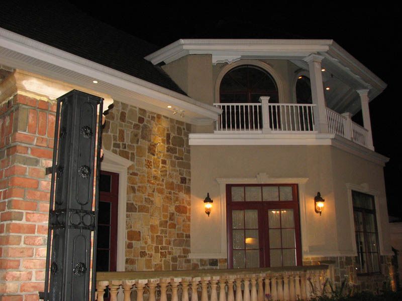 Exterior of a house at night, featuring brick, stone, and stucco with a balcony. Red door frames and sconce lighting.
