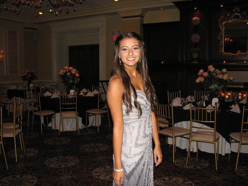 Woman in a silver beaded gown smiles in a ballroom with tables set for a formal event.