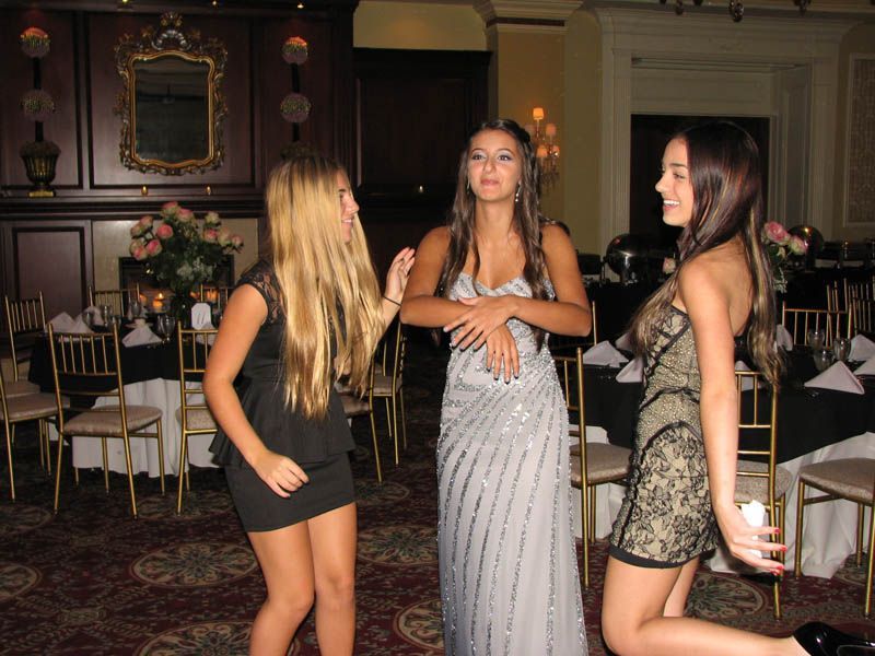 Three women in formal dresses dancing at an event, with tables and decorations in the background.