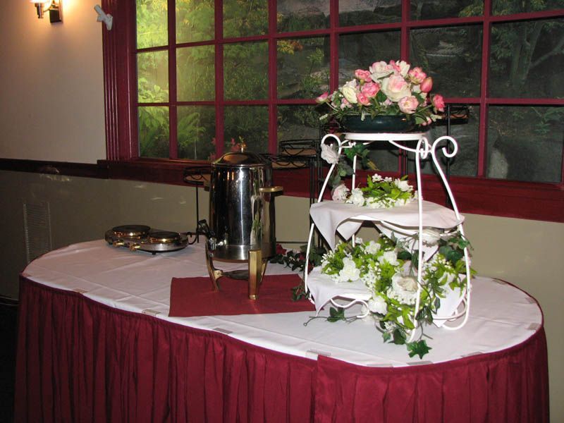 Coffee station with floral arrangement on a white tiered stand, set on a table with a maroon skirt.