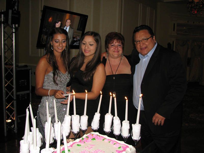 Family gathers around a lit birthday cake; two young women, a woman, and a man smile.