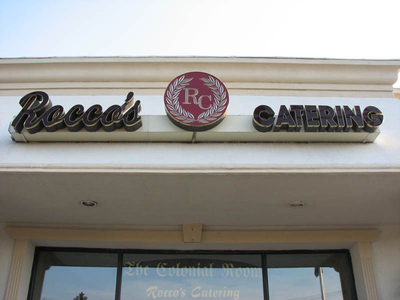 Sign for Rocco's Catering above the entrance of a building. The sign is brown and includes a logo in a red circle.