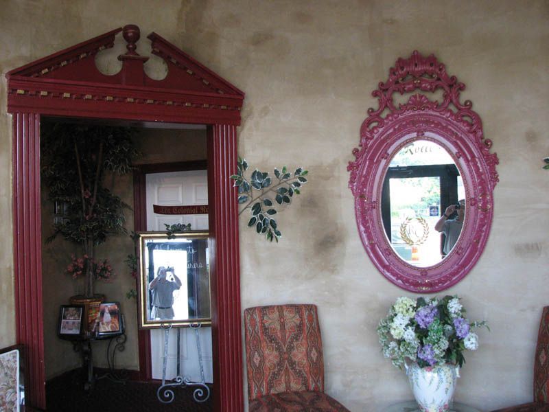 Red framed doorway and mirror, and an oval mirror on a beige wall, with a chair and flowers.