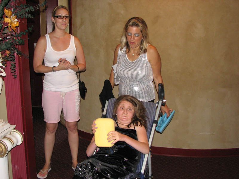 Three women inside a building. One in a wheelchair holding a yellow sponge, two others standing nearby.