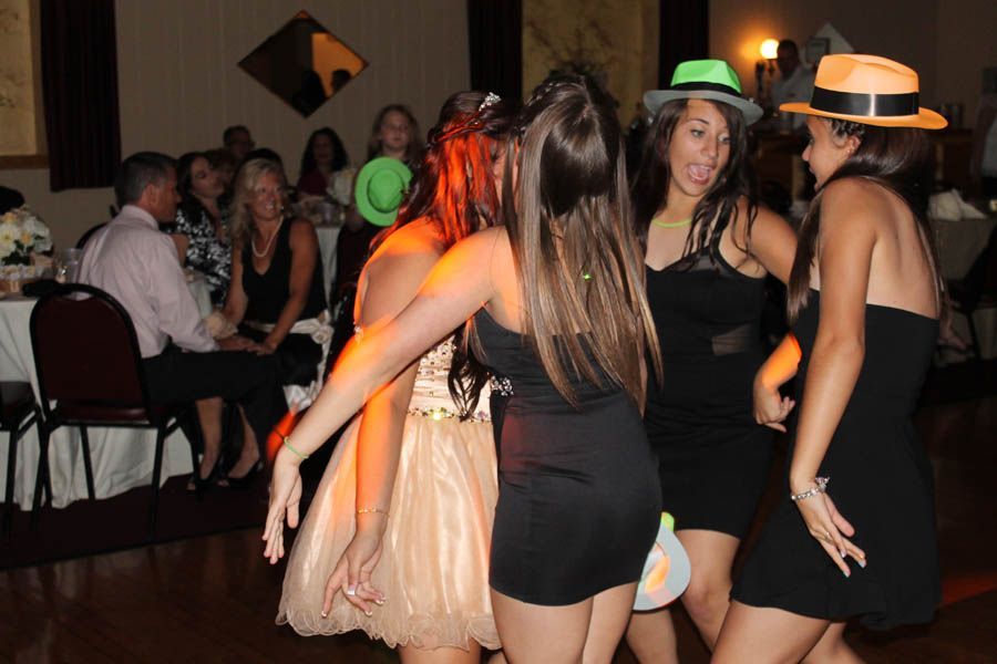 Four young people dancing at a party, wearing hats. Other guests seated at tables in the background.