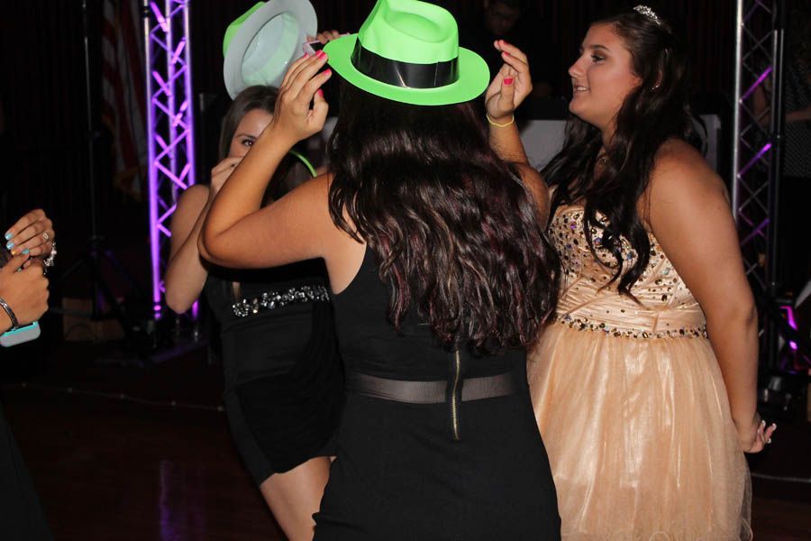 Three people dancing at a party; one woman wears a green hat.  Black and gold dresses, purple lights.