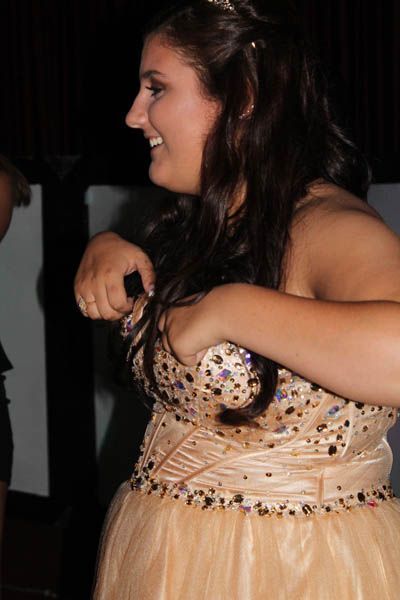 Woman in a peach-colored formal dress adjusts her chest area, smiling.  Dark hair, sparkles, and a dark background.