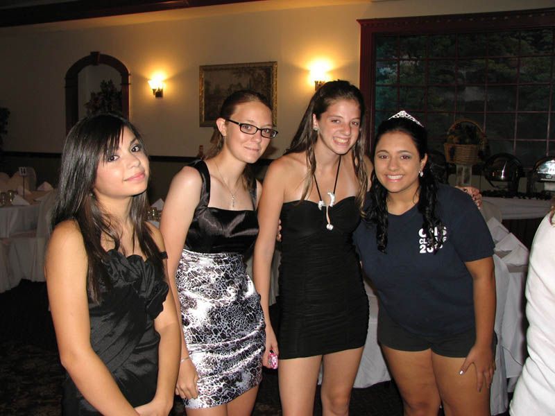 Four young people at a gathering. Three are in dresses, one in a t-shirt. Standing indoors near tables.