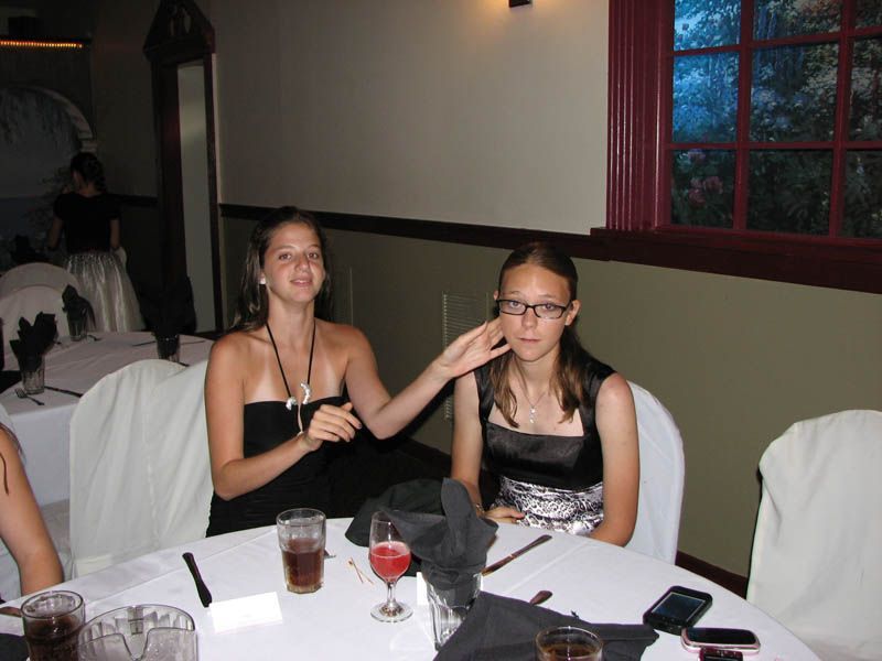 Two women at a table; one adjusts the other's face. They wear formal dresses.  Setting is a banquet hall.
