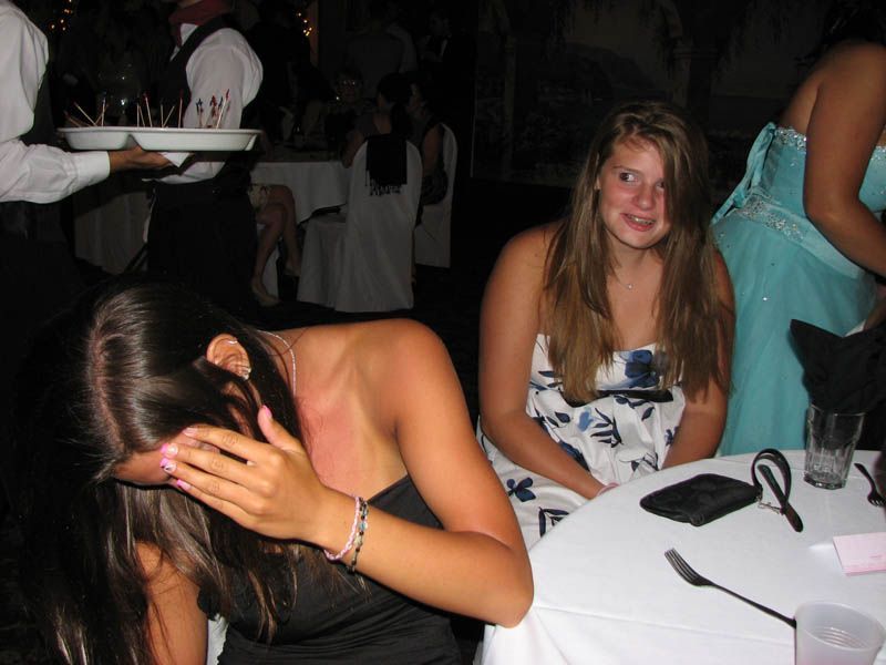 Two young women at a formal event; one covering her face, the other looking surprised. A waiter holds a tray.