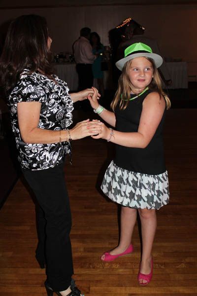 Woman and child dancing at an event; child wears a hat and patterned skirt; wooden floor.