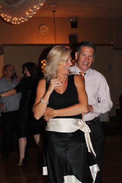 Couple dancing in a dimly lit ballroom; woman in black dress with white sash; man in pink shirt smiles.