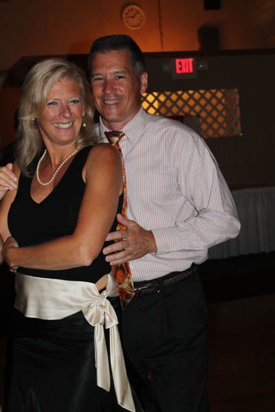 Couple dancing at an event; man's hand on woman's back, both smiling. Formal attire, dim lighting.
