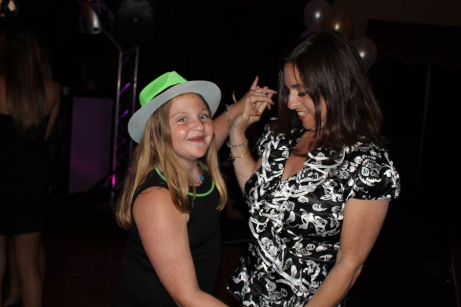 Girl in neon hat dances with a woman at a party; dark setting with balloons.
