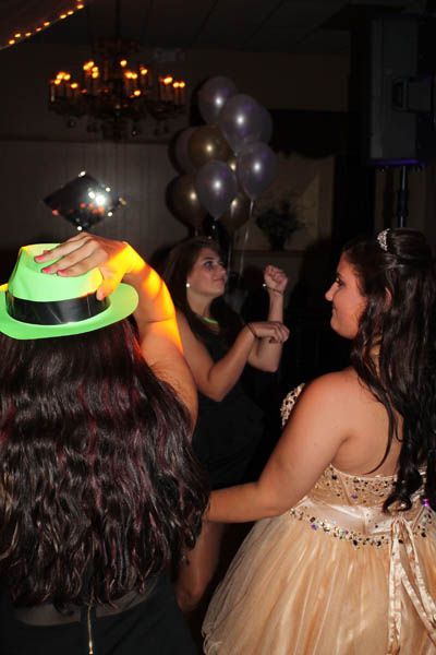 People dancing at a party; one wears a neon green hat, another a gold dress. Balloons and chandelier are visible.